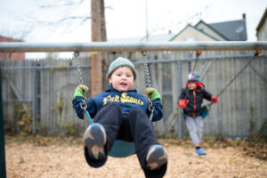 Two young children swing on swings wearing winter hats and sweatshirts