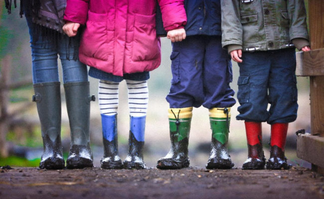 Four children, shown from the waist down, wearing coats and mud-covered rain boots