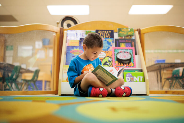 A young boy with dark hair reads a book while sitting on the floor by a bookshelf