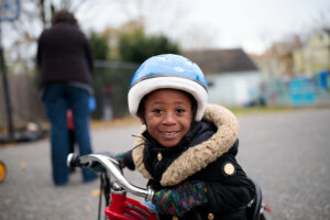 A young Black girl smiles while riding a tricycle and wearing a winter jacket and helmet