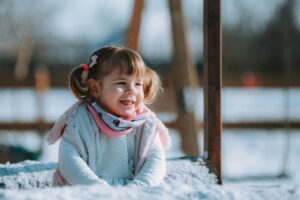 A young white girl with brown pigtails smiles while sitting in the snow