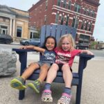 Two smiling girls sit in a chair together by the main street of Middlebury, Vermont