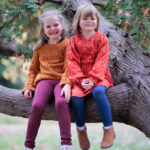 Two young blond girls with light skin sit happily in a tree together