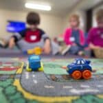 Two small plastic toy cars with eyes, on a play mat with a road printed on it, with three toddlers in the background