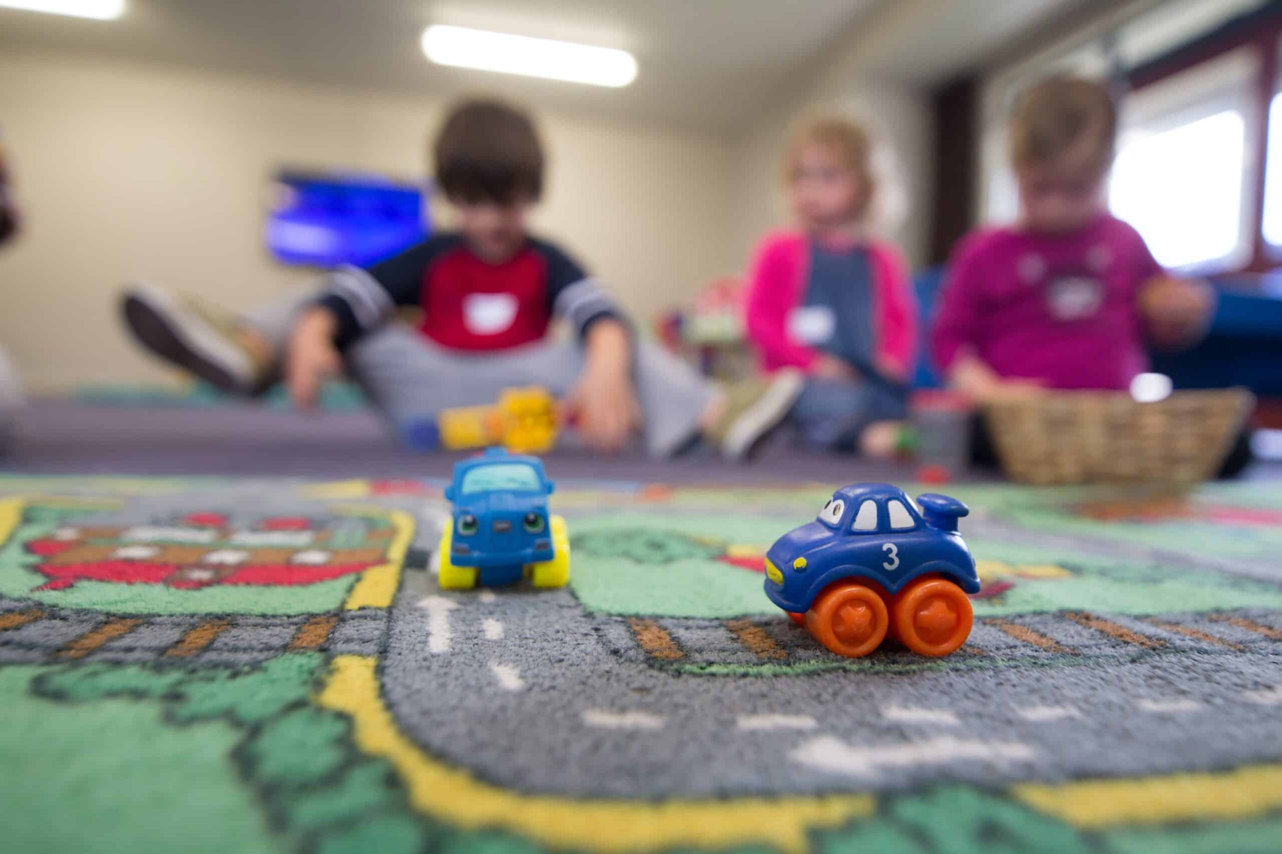 Two small plastic toy cars with eyes, on a play mat with a road printed on it, with three toddlers in the background