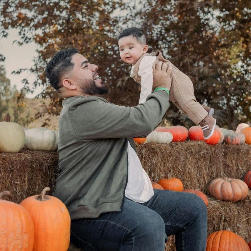 Parent lifting young child in arms with pumpkins and hay bales in background