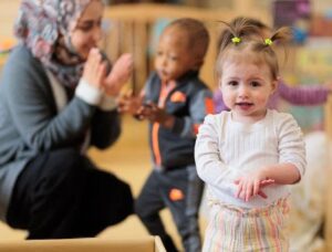 Toddler with pigtails looking at camera, with teacher in headscarf playing pattycake with another toddler in background