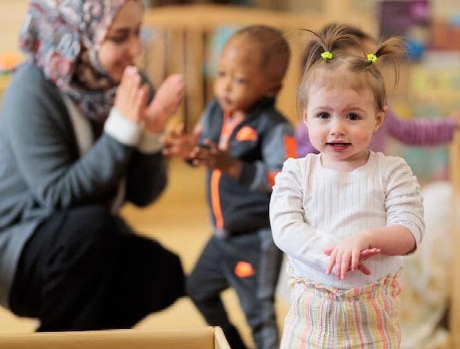 Toddler with pigtails looking at camera, with teacher in headscarf playing pattycake with another toddler in background