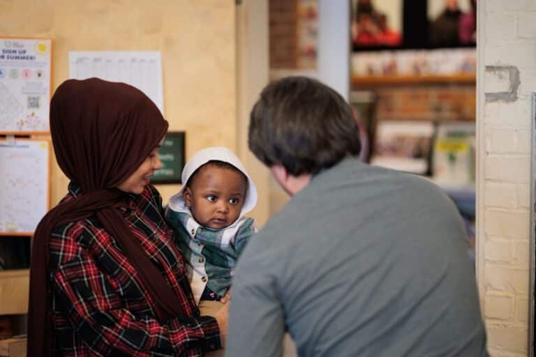 A mother wearing a head scarf holds an infant who is looking intently at another adult