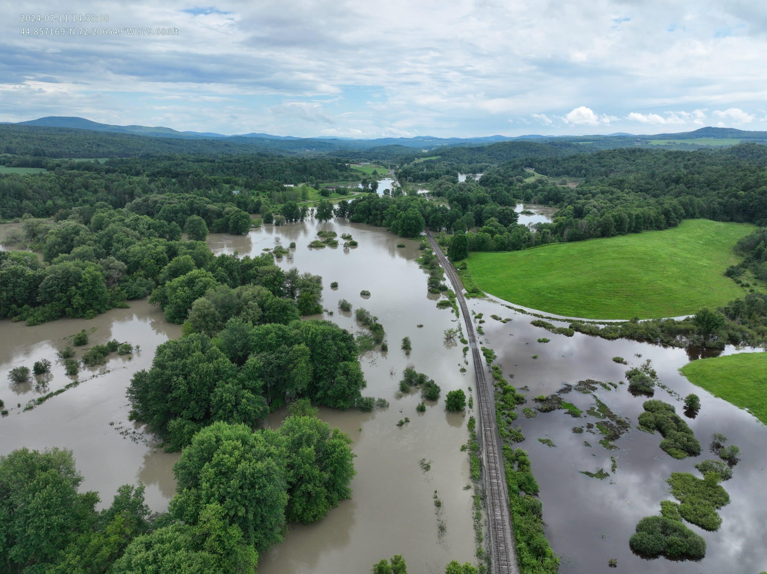 Flood waters surrounding railroad tracks and covering most of the landscape, with the tops of green trees visible above the waters and mountains in the background