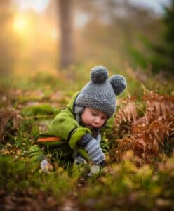 Toddler in sweater and knit hat seated and looking down, outdoors surrounded by fall leaves and ferns