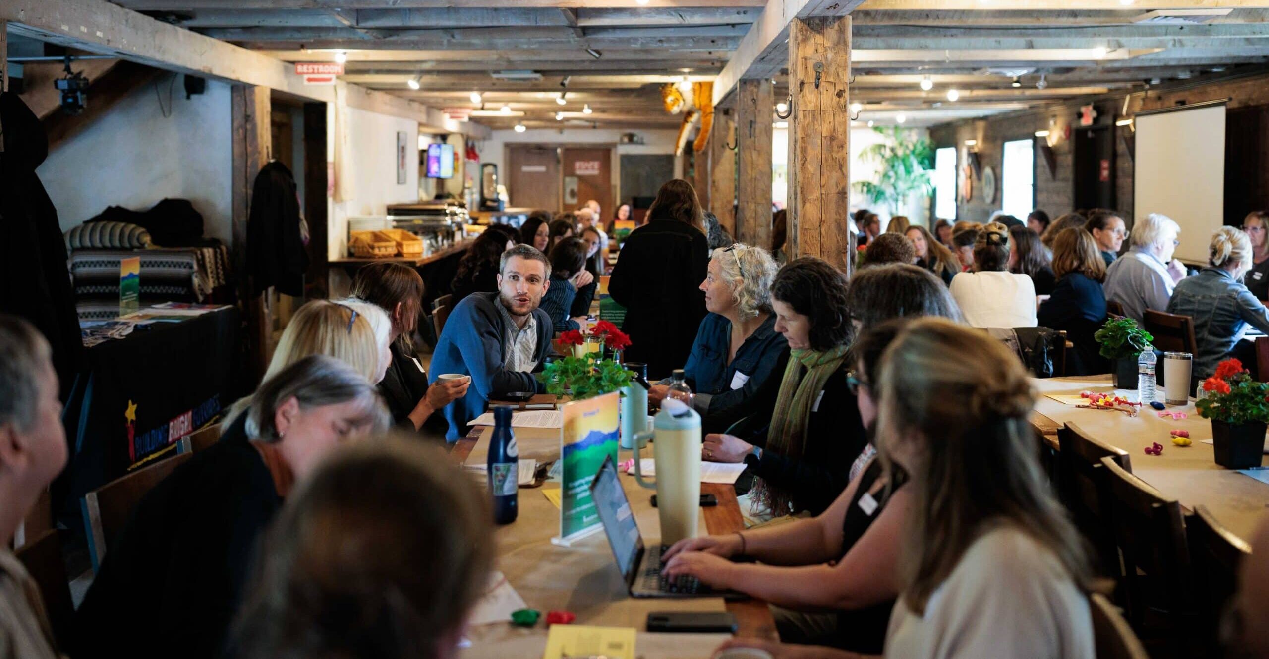 A rustic room with exposed wood beams with multiple tables of people participating in a large meeting