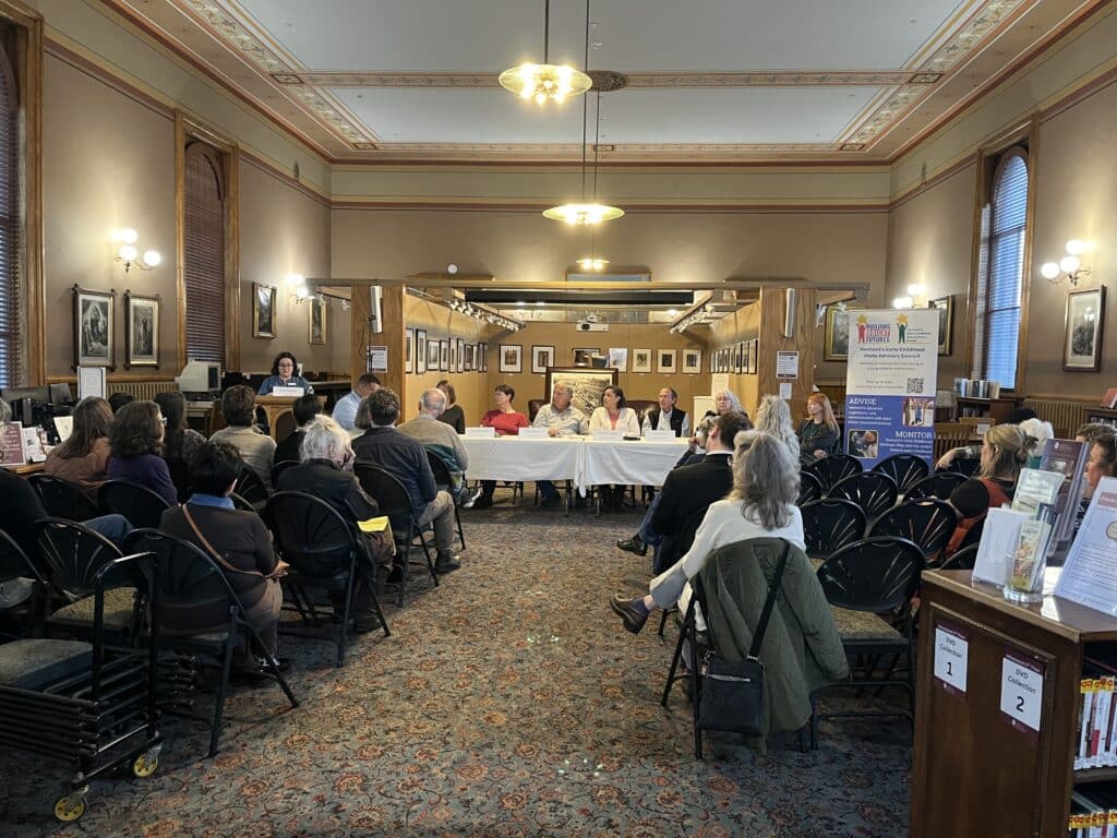 Candidates sit at a table at the front of a room while participants sit in chairs facing them