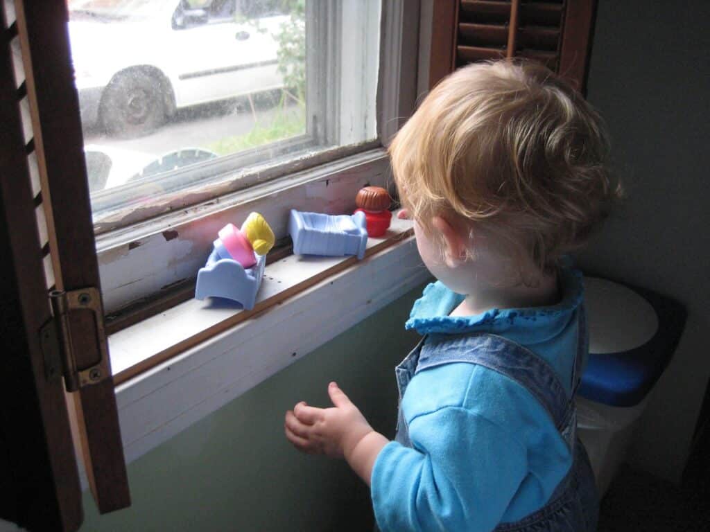 Toddler stands at window sill playing with small plastic toys. The window sill has peeling paint.