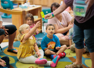 A group of young children in a child care setting, with one child reaching to take an object from a teacher
