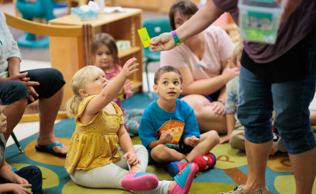 A group of young children in a child care setting, with one child reaching to take an object from a teacher